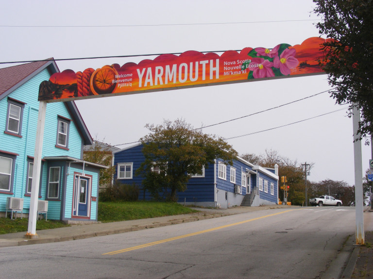 A sunset-coloured banner over a street reads "YARMOUTH." "Welcome" and "Nova Scotia" are written on either side of the banner in English, French and M'ikmaw.