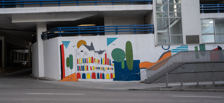 Colouful building with windows and arches in red, blue, and yellow. Simplified trees on right side in body of water with ground on the left side with trees.
