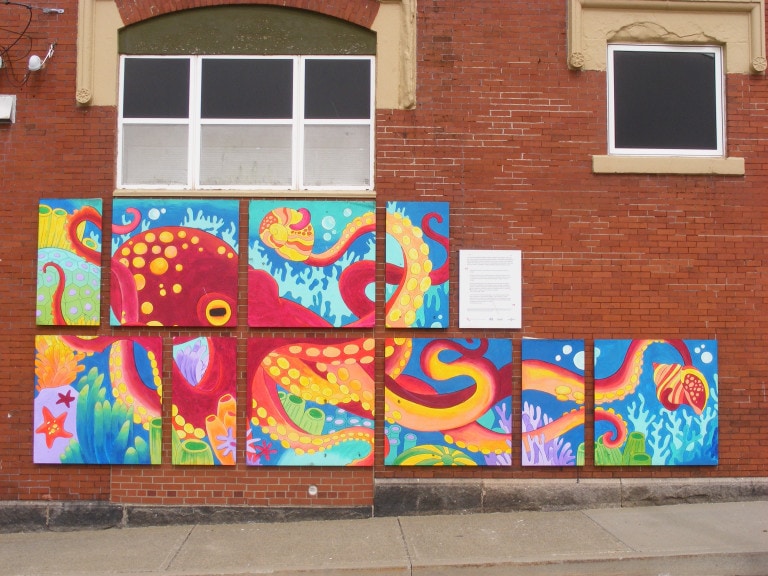 A red octopus is depicted in the ocean, surrounded by bubbles, coral, seashells and ocean vegetation. The mural is done in a serious of panels that are placed on the red brick wall of a local business.
