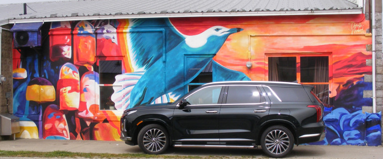 A seagull is taking flight against a vibrant, sunset-coloured background. A blue, rocky shoreline lies to the right of the seagull and a wall of buoys to the left. A black truck is parked in front of the mural.