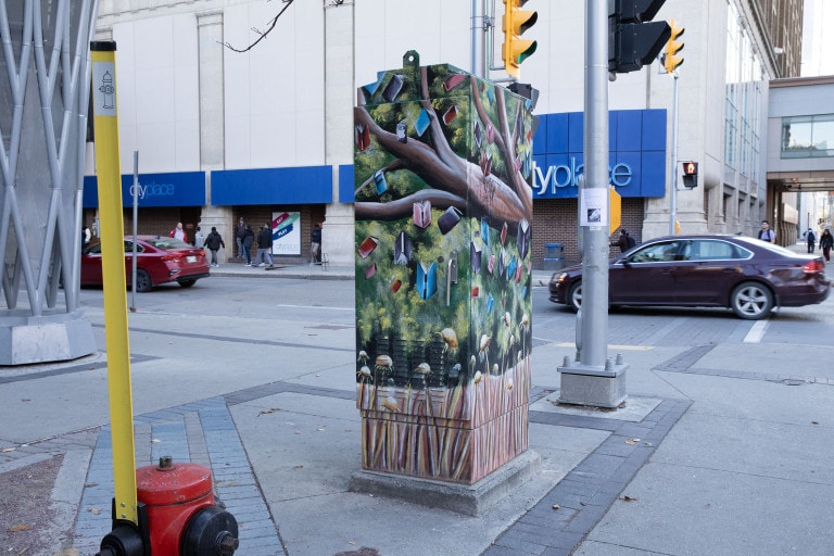 Transit box painted with a mossy, green background, with tree branches in the foreground. Books fall from the sky, towards the bottom, where yellow daisies and grass are growing.