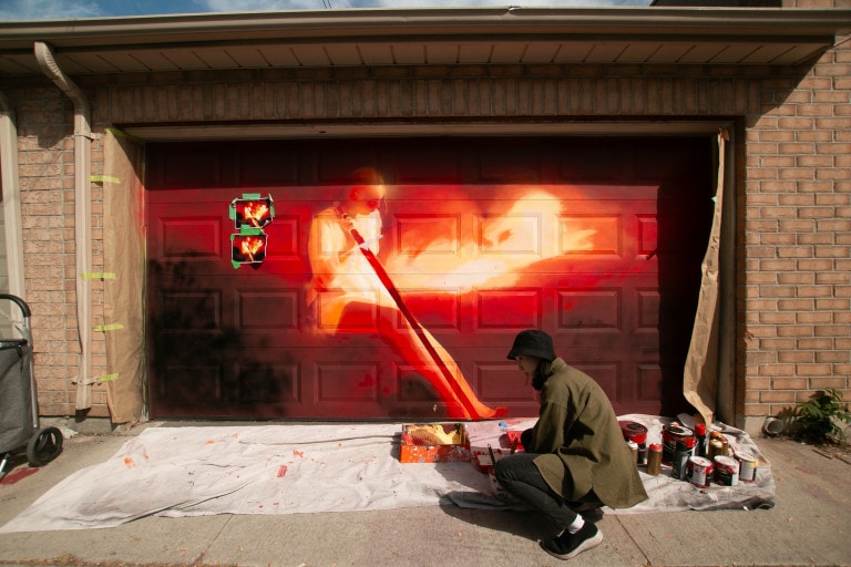 Photo of an artist at a garage door mural sitting on the floor with supplies around her. The mural is of a dark burgundy scene with a glowing yellow figure at the centre.