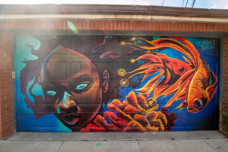 Photo of garage door mural of a female African figure with glowing blue eyes and fighter fish swimming beside her above glowing coral.