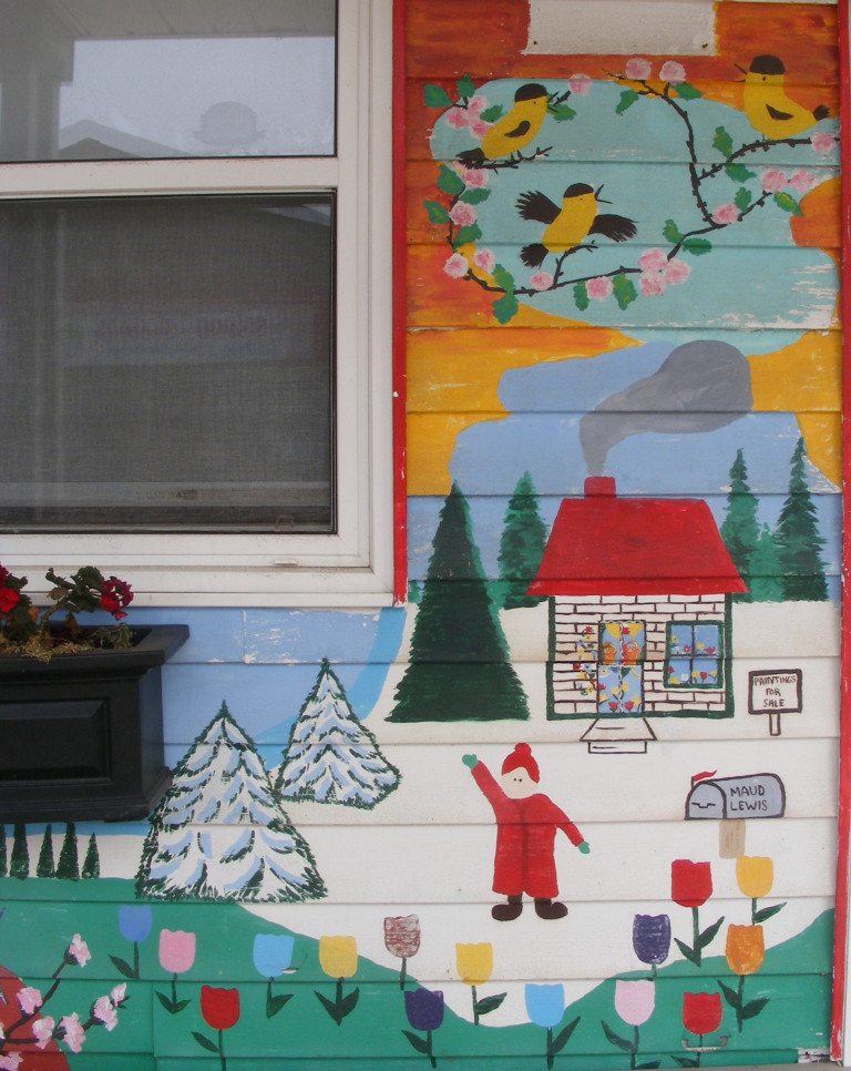The side of a building is painted with a wintery, snow-filled scene with tulips and green grass in the foreground. A figure in a red snowsuit stands in front of a white house with a red roof. The mailbox reads "Maud Lewis" and yellow birds are perched overhead.