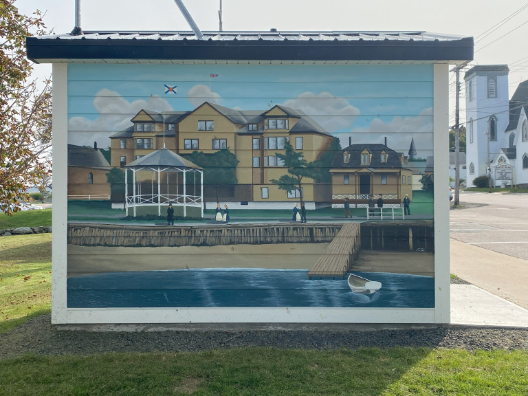 A mural is painted on the side of a small building. It depicts a shoreline scene - calm waters give way to a sandy beach and an opulent building. A gazebo and several well-dressed figures stand on the boardwalk.