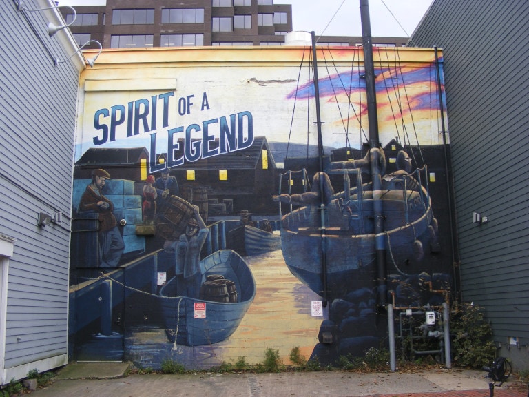A shipyard is set against the pale sunset. A man in a boat carries a barrel over his shoulder. There are some men and a woman in a flapper dress standing on the dock. A sailboat is off to the side. "Spirit of a Legend" is written in bold on the top left corner.