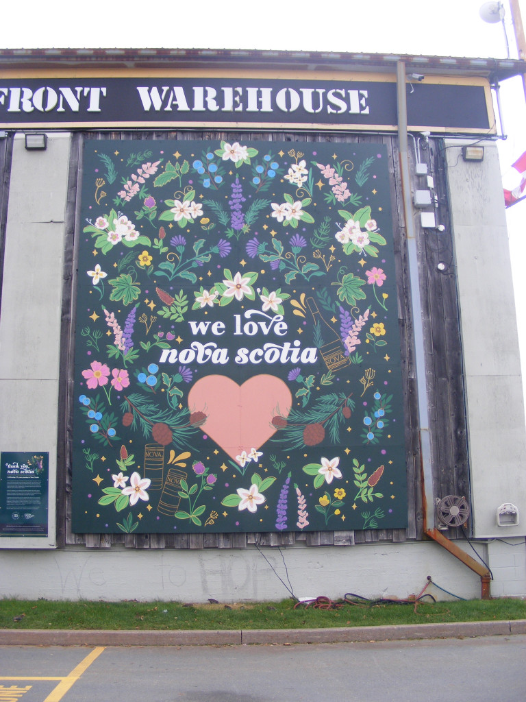 White lettering reads "We Love Nova Scotia." The lettering is surrounded by berries, flowers, cones, branches, and cans and bottles of NOVA 7 wine.