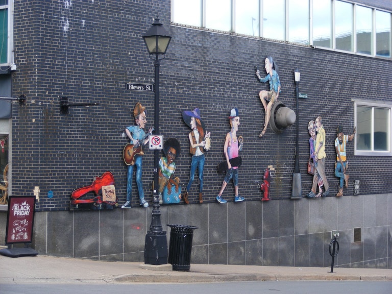 A cast of characters are painted on a wall: a man with a guitar, a man crouched down with a suitcase, a woman with a tambourine, a man with a skateboard, a man in an orange vest and an elderly couple.