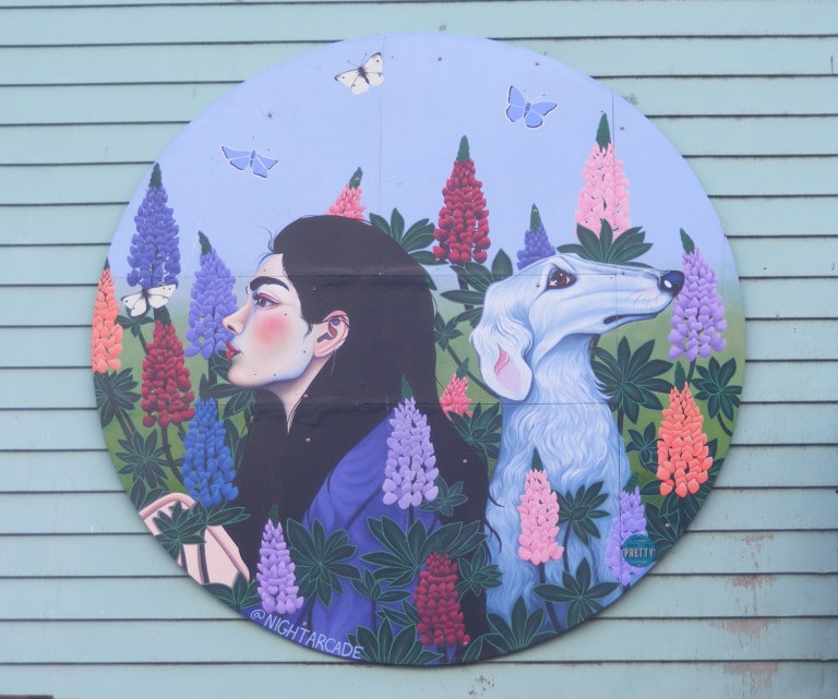 A woman sits back to back with a white, long-haired dog. Lupins and butterflies encircle them.