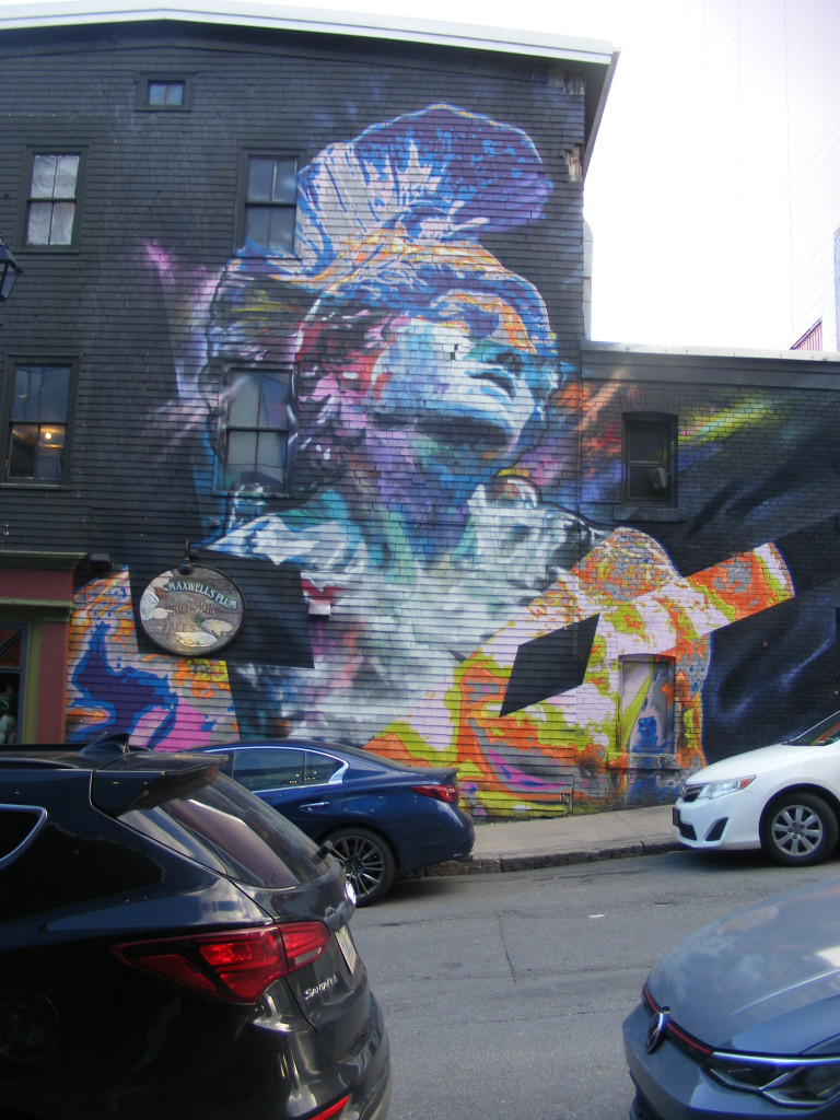 A Roman gladiator, glowing blue, purple, pink and orange is set against a black background on the side a building. The gladiator's head is tilted back.