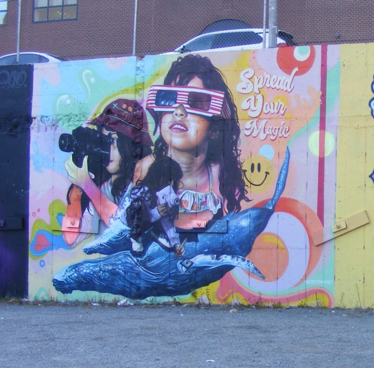 Three young girls form the center of this mural. One is looking through a camcorder, one is looking forward with 3D glasses and one is sketching with one hand and holding a Polaroid to her eye in the other. They're resting on two blue whales. The background is pastel swirls. "Spread your magic" is written next to the girl with 3D glasses.