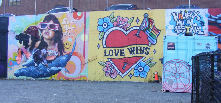 A red heart, painted in the style of an American traditional tattoo,  holds a banner reading "Love Wins." Flowers and a LGBTQIA+ flag frame the heart. There's another mural located to the left and lettering that reads "Halifax Mural Festival 23" to the right.
