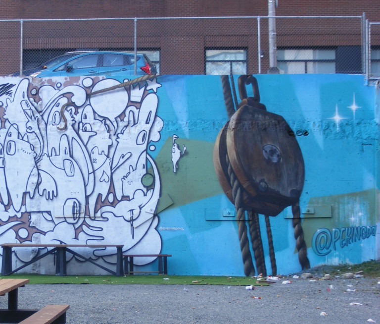 A rustic pulley is painted on the wall with a blue background. The artist's signature is visible: @pekingdon. There's another mural on the left.