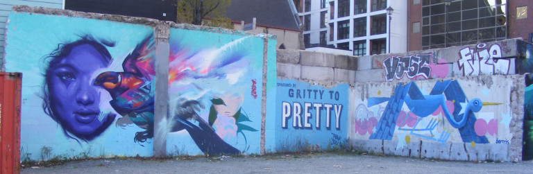 A woman's face is painted with shades of blue. She stares forward. A warm-toned bird shadows half of her face. To the right of the mural is the "Gritty to Pretty" logo.