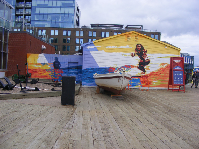 The side of a building is painted with bright, sunset colours and blue ocean waves. A woman faces forward, stretching her arms outwards on a surfboard. A woman to the left sits on her surfboard, her back facing the viewer. There's a boat in front of the mural.
