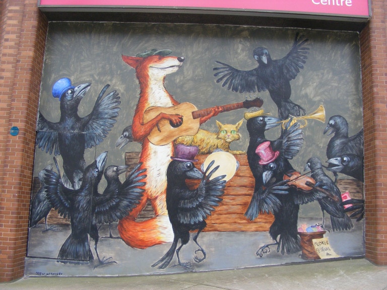 Crows in colourful top hats dance, play the fiddle and play the tambourine surrounding a fox playing the guitar.  They dance behind a a tip jar that reads "Busker Festival"