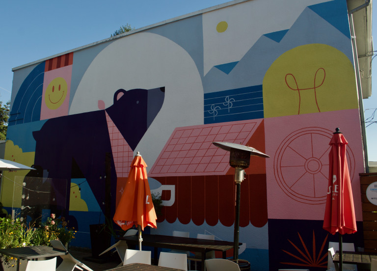 A vertical wall mural, with colourful geometric shapes of a natural landscape with a bear in the foreground walking towards a pink house