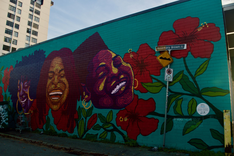 A vertical wall mural with a teal background depicting three women laughing surrounded by flowers.