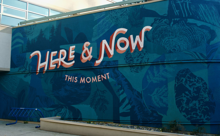 A blue vertical mural background with white letter and red drop shadow that reads Here & Now this moment