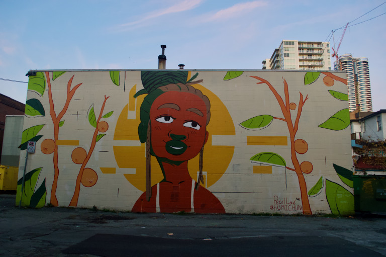The mural depicts a smiling Black woman looking off into the distance, surrounded by a warm background that includes fruit trees and a sun emanating from behind her.
