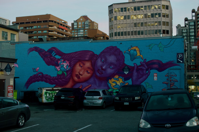 A trans/non-binary couple embrace each other in a dewy, whimsical scene featuring flowers, mushrooms, and other elements of the natural world.