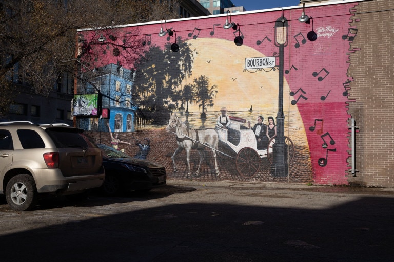 A man and woman couple wear formal attire on a carriage, being pulled by a white horse. Another man in a bowtie and hat sits at the front of the carriage, smiling towards the viewer. To the left of this, is a female dancer and a man who plays a clarinet. This occurs infront of a sunset, that sets above a body of water. There is a blue building to the left, and a sign that reads "Bourbon St." to the right.