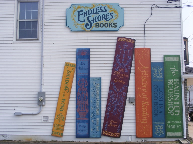 The side of a building is decorated with the spines of a collection of books. The sign above the mural reads "Endless Shores Books." The spines are all different colours and feature ornate piping and titles such as "A History of Reading" and "The Haunted Bookshop"