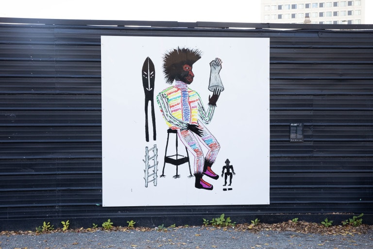 Man sits on a black stool wearing a brightly coloured shirt and pants with stripes. He puts his hand into a light blue glove. Next to him is a blue wooden ladder and a mask-like shape. Below his feet is a small black silhouette figure.