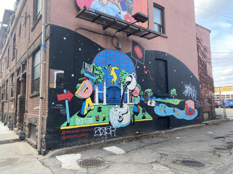 Photo of an illustrative mural on black background featuring a surrealistic image of a head laying down with coffee poured into his mouth. The background has a rabbit it a beer bottle, Trinity Bellwoods gate, a laptop, large letters “D” & “A”, and dogs playing, among other elements. Various business names are featured in the artwork, including Tokyo Smoke, Design Agency, Flow and The Fueling Station.