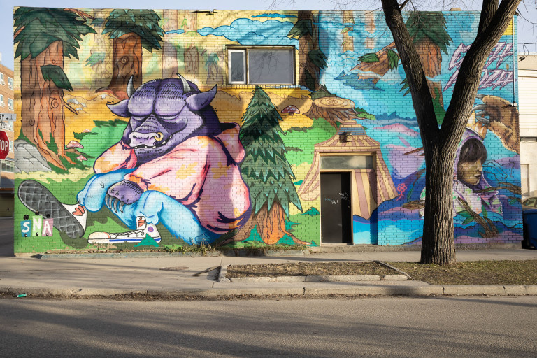 A purple bull is sits wearing a bright sweater and blue jeans. He sits in a bright forest. To the right is a woman in a purple garment who gets a crown put on her head by an animal.