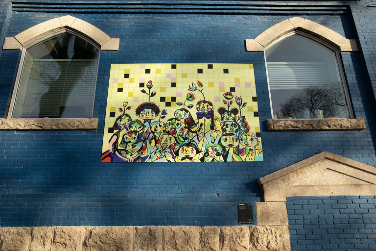 On a tile background, several sketched figures with bright coloured shapes on their faces stand in a group. Flowers grow behind them.