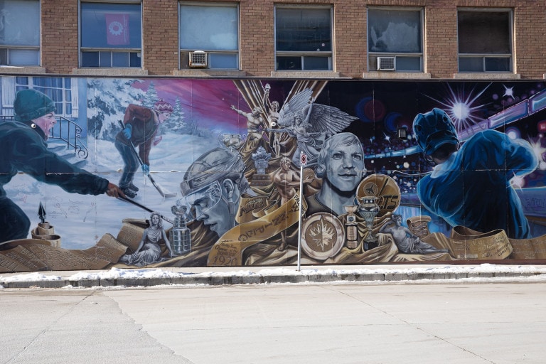 On the left, two children are seen yelling, playing hockey on an outdoor rink. The middle of the mural displays a collage-like arrangement of golden and silver hockey trophies, symbols and players. To the right, a professional NHL player looks out at an arena crowd, while swinging his stick.