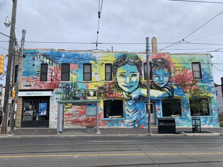 Photo of a a large scale mural on the side of a building featuring linework portrait of a female with a boy on her back and the Toronto city scene beside her with a street car at the focal point. The entire background underneath the linework is painted in red, blues and yellows in what mimics a water colour painting.