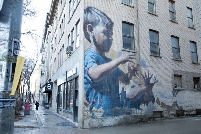 A young boy in a blue shirts stands with his hands in front of him, and fingers spread out. His hands are repeated just below him. Various grey and brown shapes surround him.