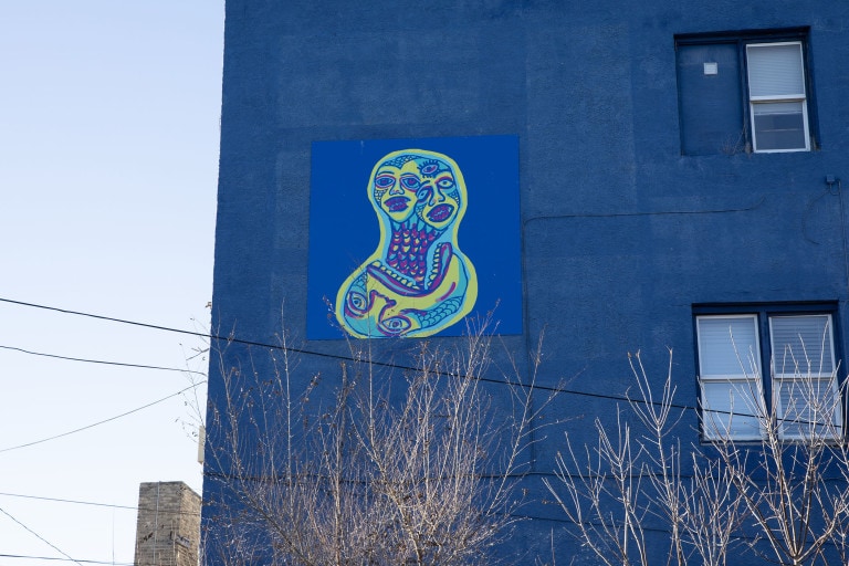 On a blue panel background, a two headed figure emerges from another figure's mouth. Each figure is green, light blue and magenta with scales on its face and neck.