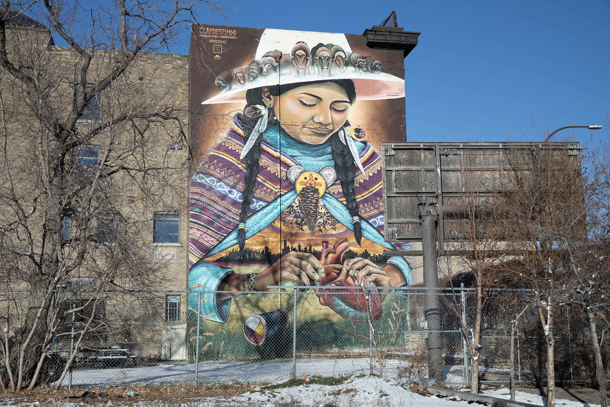 A woman with dark hair in braids looks down to the human heart that she sews with thread. The bobbin has the medicine wheel symbol on it. The woman wears a white hat with a line of muskox on the brim and a shawl, with a landscape scene painted beneath it. The shawl has a pinecone in the centre of it.