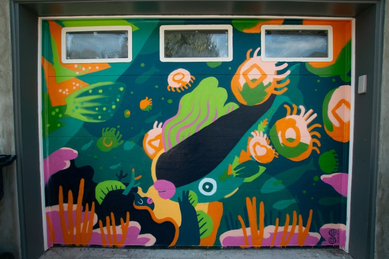Garage door mural of an underwater scene in shades of blue and green colour blocks. The main subject is a fish with pink lips, swimming downwards towards a figure holding it in what appears to be an embrace. Jellyfish and other aquatic creatures swim around it.