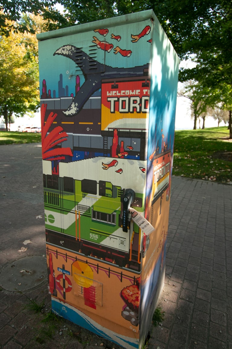Photo of a utility box with a digital art wrap featuring pixelated art. This piece features the Go train at the centre, a highway above it with a “welcome to Toronto” sign on the side.