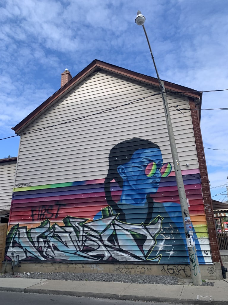 Photo of a mural on the side of a house of a blue-skinned woman with braids and round sunglasses on the right and looking to the right. Graffiti lettering is below her and on the background are psychedelic colours of the sky that reflects in her sunglasses.