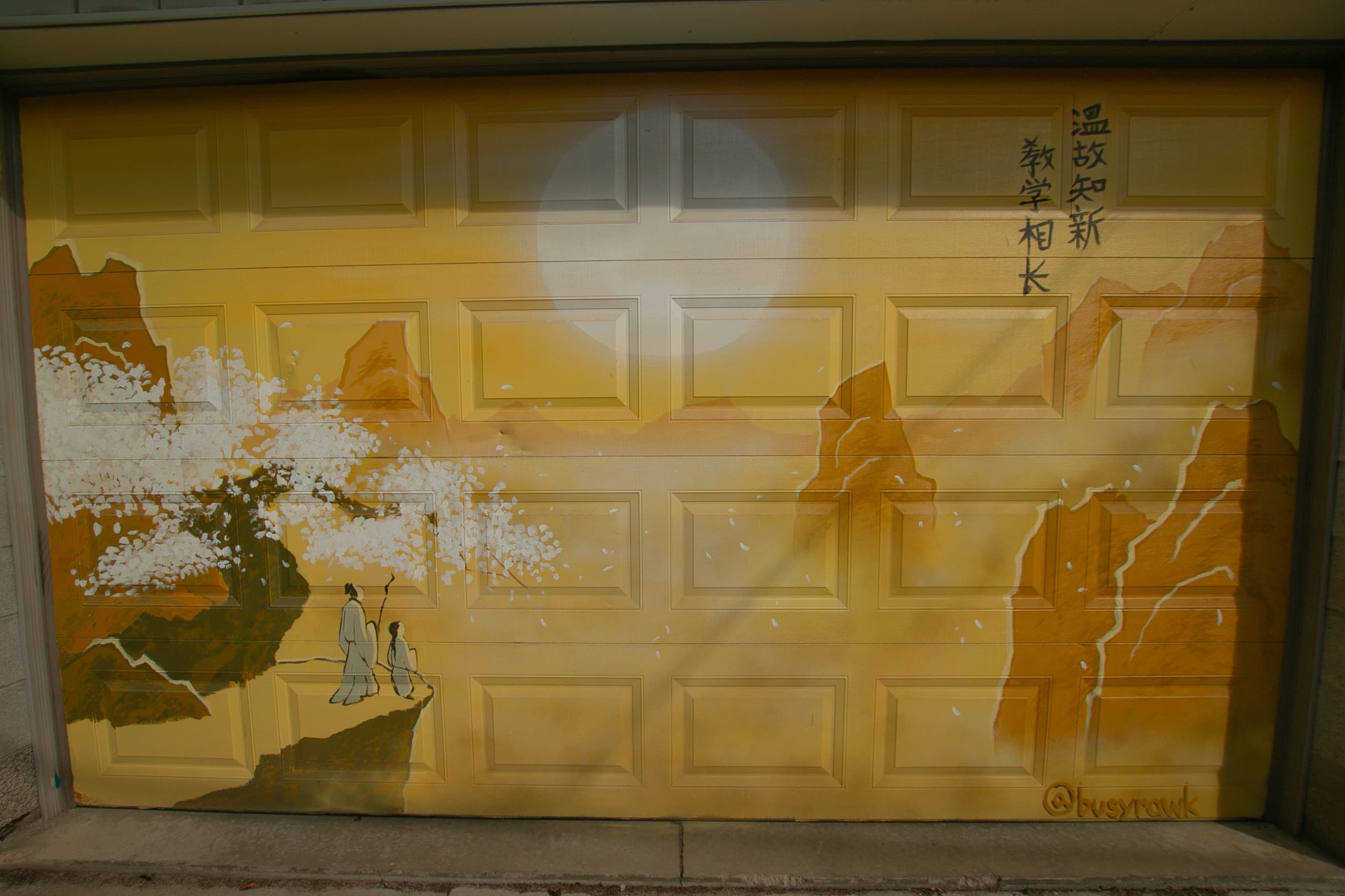 Photo of a garage door mural in yellow of a Chinese landscape scene of mountains in a mist and a full moon at the centre. Two characters in traditional Chinese clothing stand at the cliff and look towards the moon.
