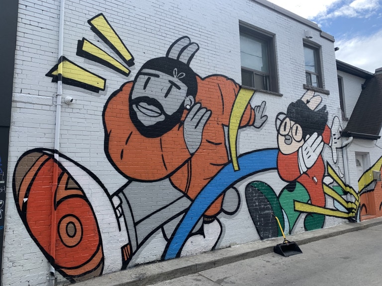 Photo of a section of a large scale wall mural with a white background and characters in the signature pose created by Chairman Ting. First character appears to be Drake, second character unknown. Comic-like motifs decorate around the characters.