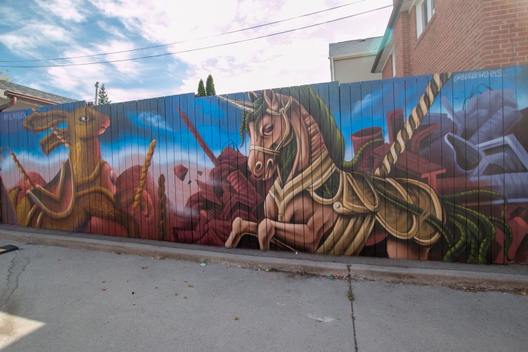 Photo of a mural on a fence featuring a rabbit and a horse in a carousel theme with an apocalyptic background of a blue sky and brown ruins.