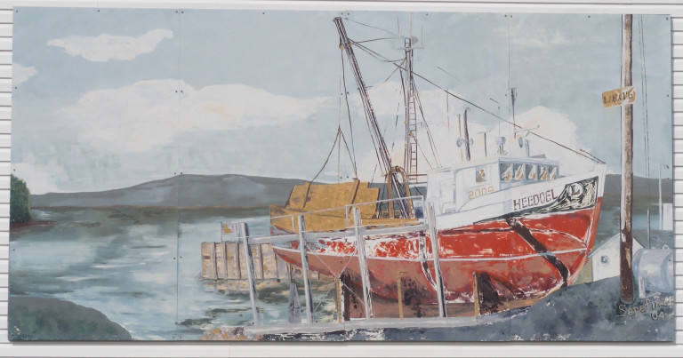 A red and white fishing boat is perched on the shore, out of the water and secured by a skid. The ocean, sky and a mass of land lay behind the boat. The boat's name is "Heedoel"