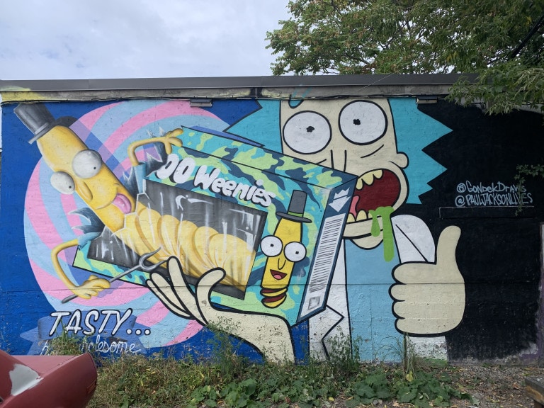 Photo of a mural of Rick from “Rick and Marty” holding a box of 00 Weenies (appears to be some sort of potato chip) with the Weenie character coming out of the box. Rick is drooling and holding a thumbs up. At the bottom right writes “Tasty… butt wholesome”