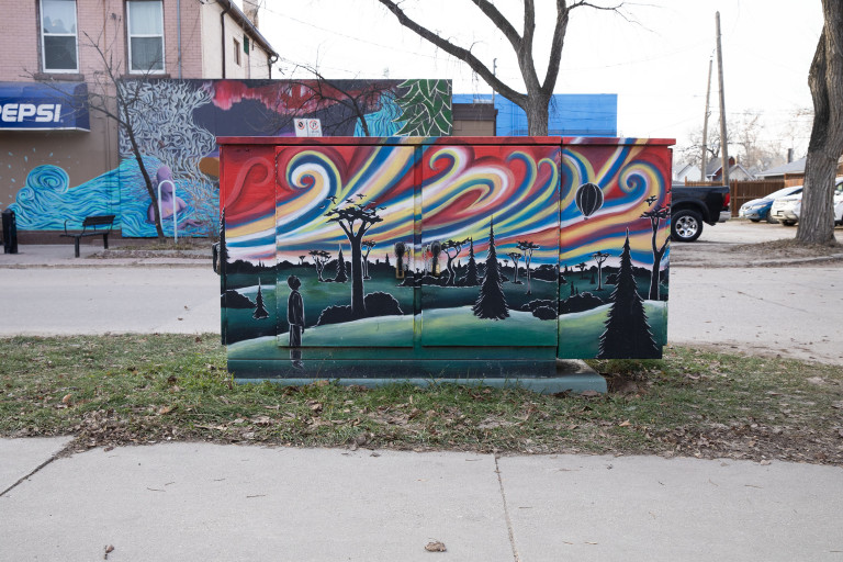 A black silhouette figure looks up at a bright coloured sky filled with vibrance and motion. The trees and bushes in the distance are also black, and sit on dark green grass.