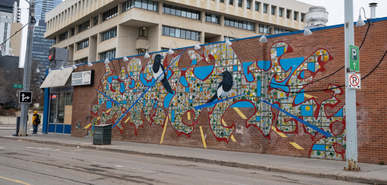 Two magpies seperated, perched on a sharp blue needle-like shape with grid of city in grey and different coloured dots.