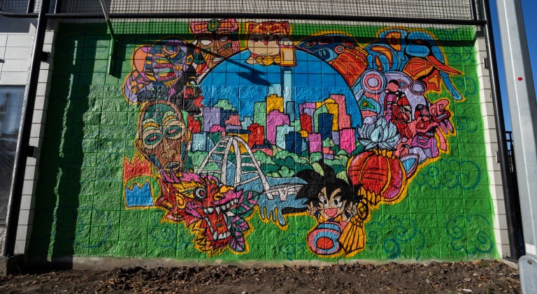 City skyline and bridge of Edmonton surrounded in colourful tribal masks, Dragon Ball Z character (Goku), and a lucky cat holding a lantern on the center top of wall.