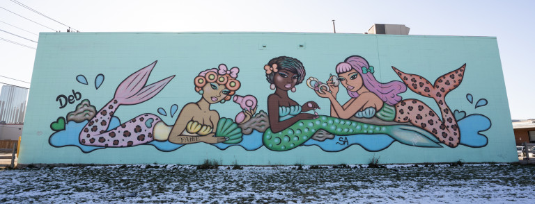 3 mermaids lying around, left mermaid holding hair dryer, middle mermaid putting nail polish on, right mermaid is putting makeup on.