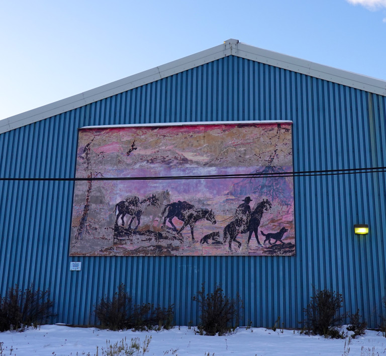 A fading and peeling mural created as part of the 1992 Yukon Sourdough Rendezvous depicts a surveyor leading a packtrain of horses through the Yukon wilderness.