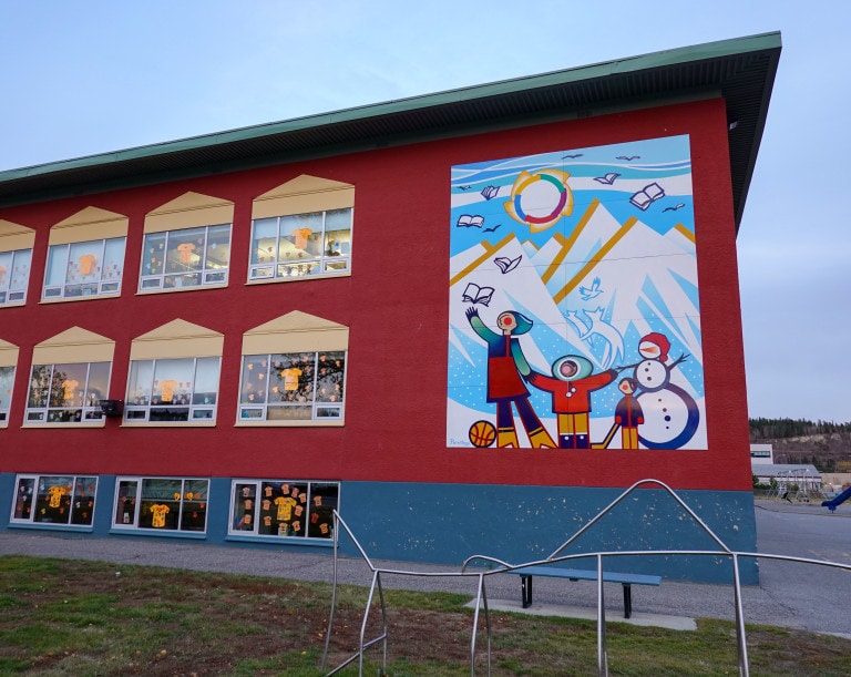 Mural of children and a snowman in front of mountain where books fly like birds on the side of a grade school where there are several orange shirts in classroom windows